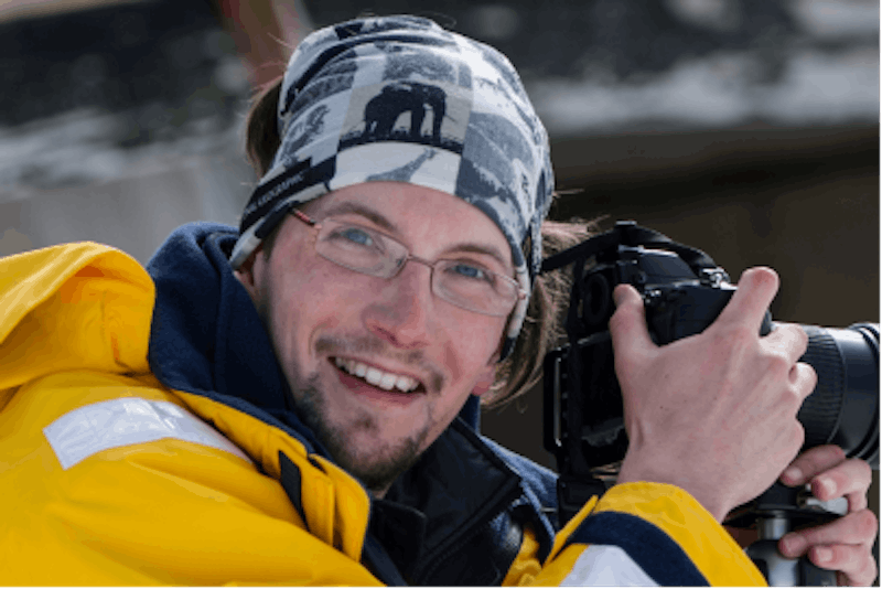 Glenn is wearing glasses, a bandanna and yellow raincoat. He is holding a camera and smiling.